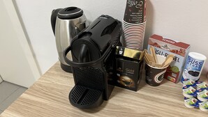A coffee maker, a stack of disposable coffee cups, a box of coffee pods, a thermos, and a bag of wooden spoons on a wooden table. - Apart Rom Hannover Centrum (Hannover)