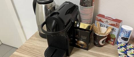 A coffee maker, a stack of disposable coffee cups, a box of coffee pods, a thermos, and a bag of wooden spoons on a wooden table.