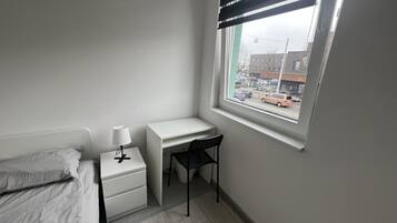 A small, neatly arranged bedroom with a single bed, a bedside table with a lamp, a desk, and a chair. A window offers a view of the street outside.