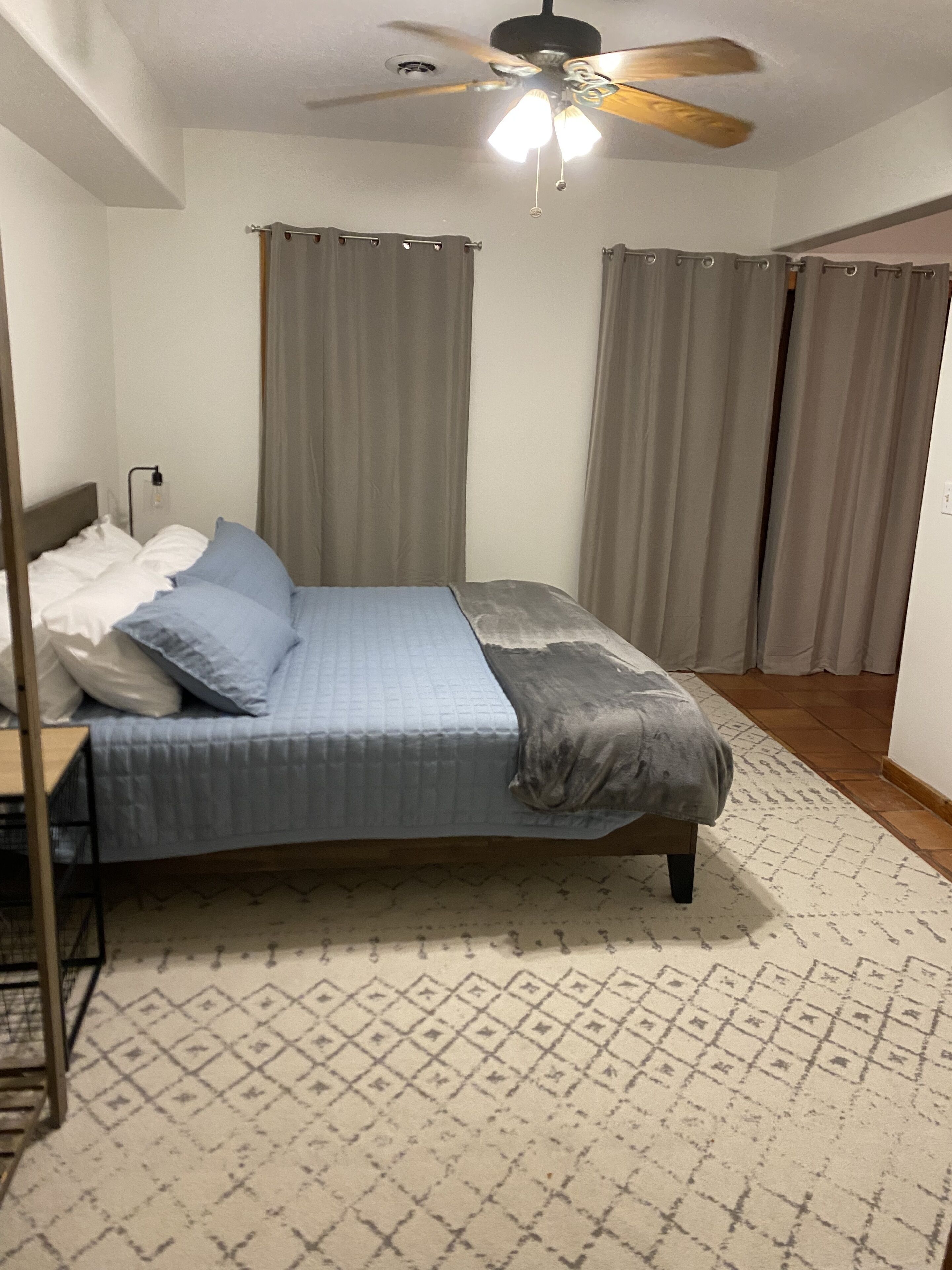 Un dormitorio con cama, cortinas, ventilador de techo y alfombra estampada.