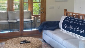 A cozy living room with a light blue sofa, a woven ottoman, and a small round table. A large window offers a view of greenery outside.