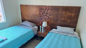 Duas camas de solteiro com roupa de cama azul combinando e cabeceiras de madeira, um criado-mudo com abajur e uma janela com luz natural.