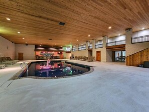 Uma grande piscina interna com um flutuador de flamingo rosa, cadeiras de descanso e um bar ao fundo.
