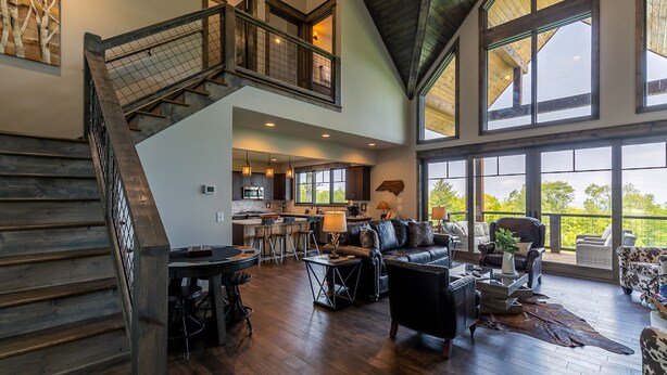 A spacious living area with a high ceiling, a staircase, a kitchen, and large windows offering a view of the outdoors.