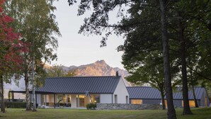 Une maison moderne avec des fondations en pierre et un toit en métal, entourée d’arbres et avec une montagne en arrière-plan.