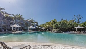 A pool area with lounge chairs and umbrellas, surrounded by palm trees and a building with balconies.
