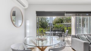 A modern living room with a glass dining table, white chairs, a grey sofa, and a large sliding glass door leading to an outdoor area with greenery.