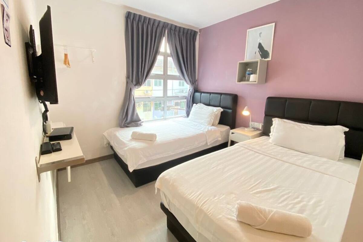 A hotel room with two beds, a flat-screen TV mounted on the wall, a window with curtains, and a small bedside table with a lamp.
