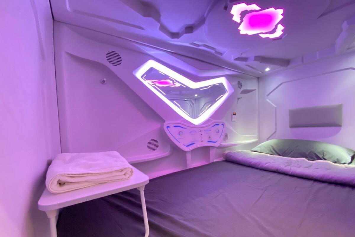 A bedroom with a bed, a small bedside table with a towel, and a neon heart-shaped light fixture on the wall.