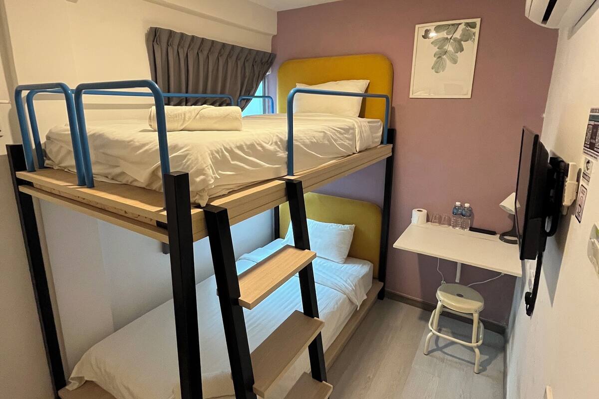 A bunk bed room with a desk and chair, a small stool, and a wall-mounted TV.