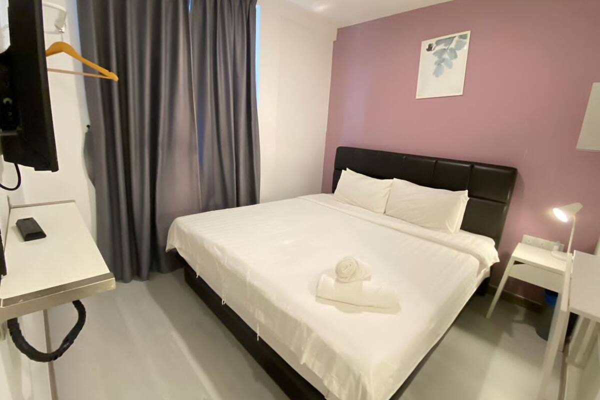 A neatly made bed with a white comforter and pillows, a bedside table with a lamp, a wall-mounted picture, and a hanging coat rack.