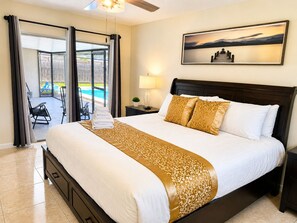 A hotel room with a bed, a nightstand, a lamp, a painting, and a view of a pool and outdoor seating.