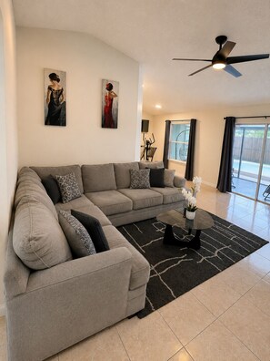 A modern living room with a sectional sofa, a ceiling fan, artwork on the walls, and a view to the outside.