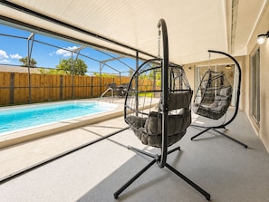 A poolside area with a hanging chair and a table, a wooden fence, and a clear view of the sky.