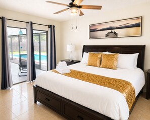 A hotel room with a bed, a nightstand, a lamp, a ceiling fan, a window with curtains, and a view of a swimming pool.