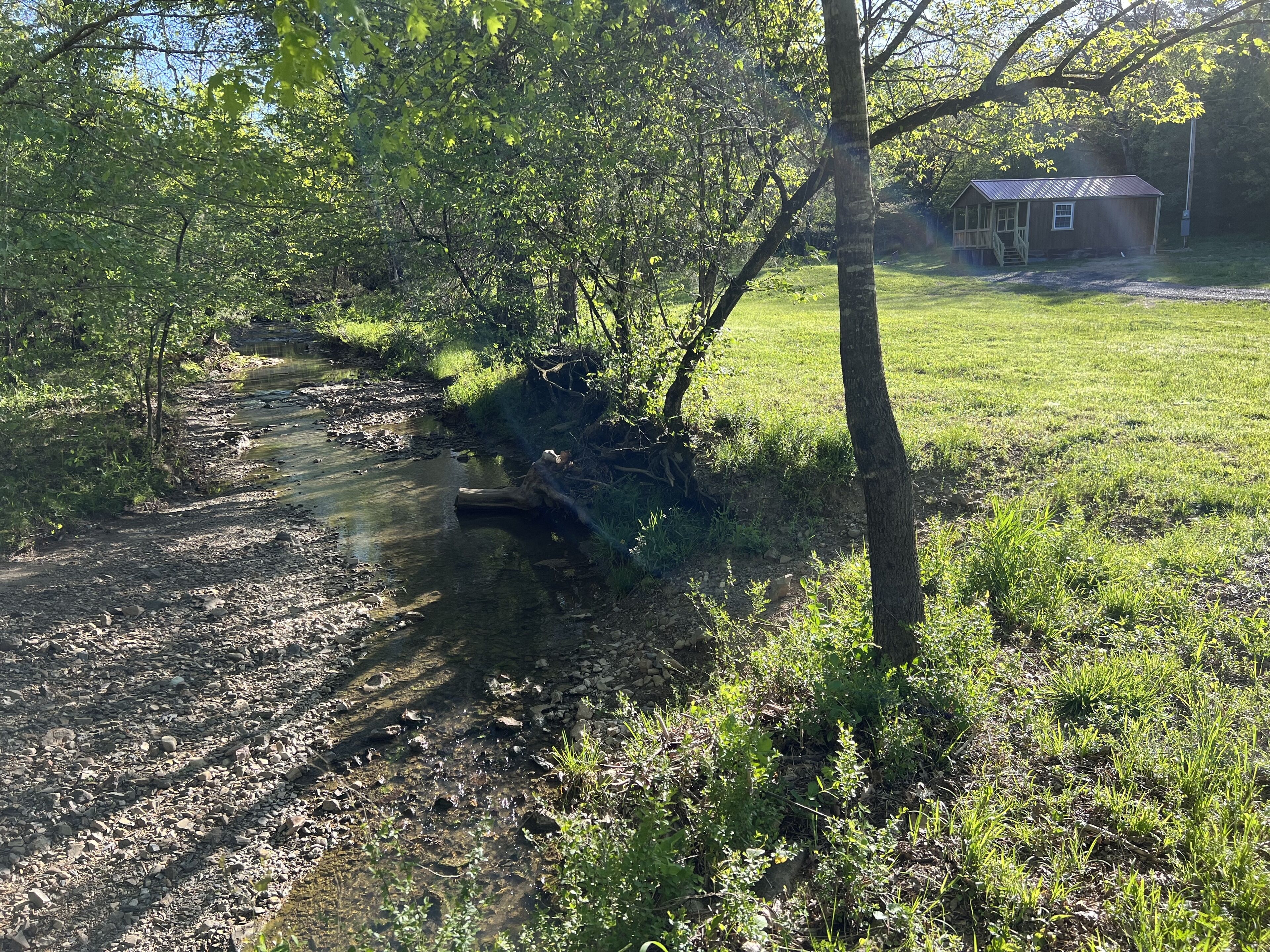 A rural landscape with a small wooden structure near a stream, surrounded by trees and grass.