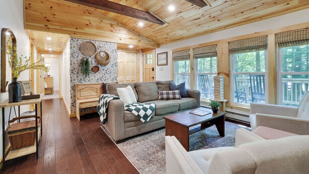 A cozy living room with a wooden ceiling, a sofa, a coffee table, and a view of the outdoors through large windows.
