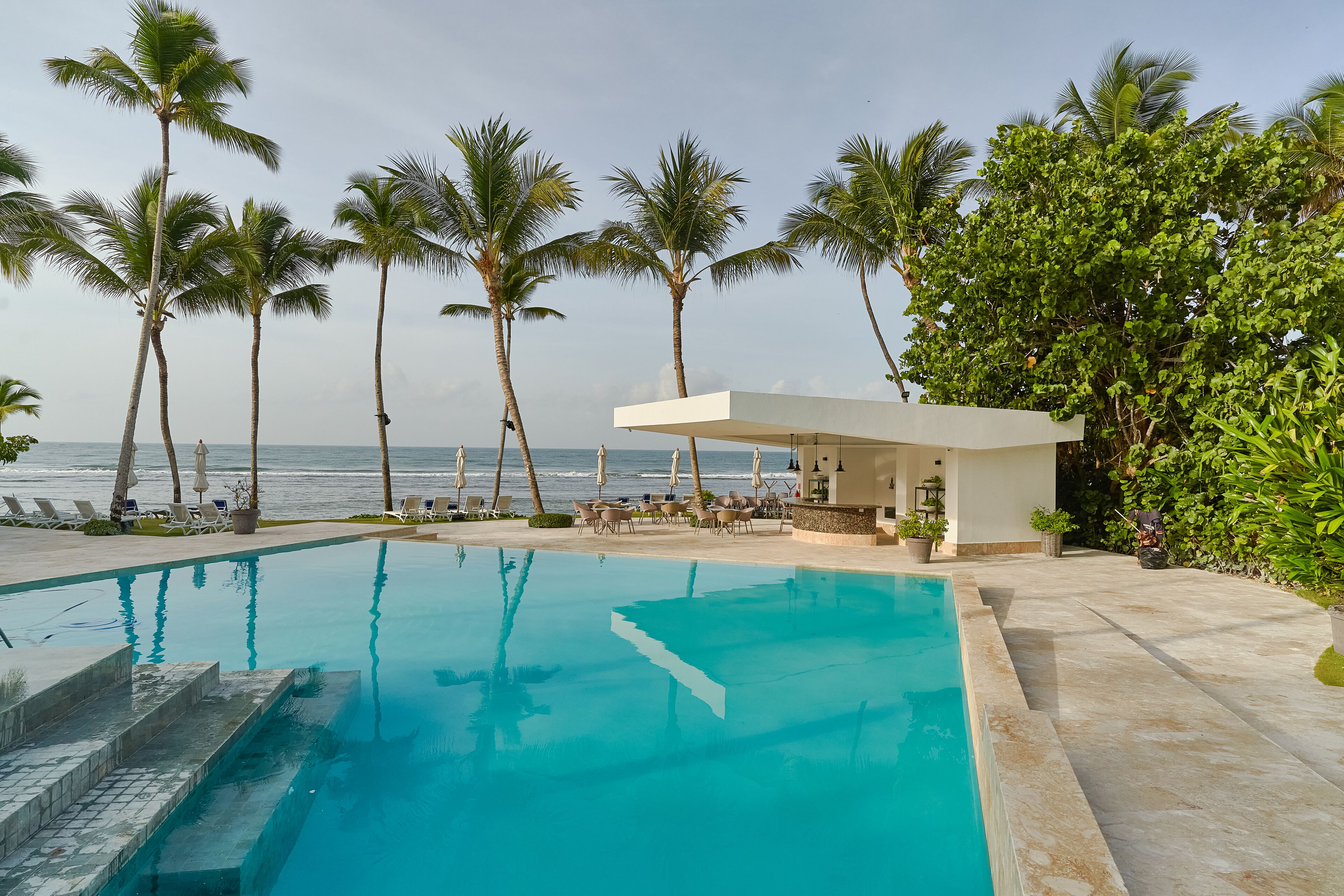 A pool area with a view of the ocean, surrounded by palm trees and a covered seating area.