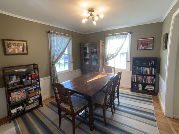 A dining room with a wooden table and chairs, a bookshelf, and a window with patterned curtains.