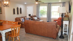 A living room with a brown leather sofa, wooden dining table, and chairs. There is a ceiling fan, a window with curtains, and a television mounted on the wall. - Gainesboro, TN House, Dale Hollow Lake, Standing Stone State Park, Roaring River (Gainesboro)
