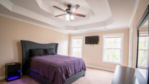 Un dormitorio con cama, ventilador de techo, televisor montado en la pared y ventanas.