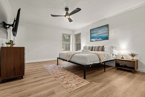 A bedroom with a bed, bedside table, nightstand, ceiling fan, and a painting on the wall.