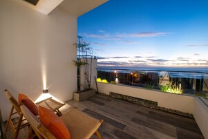 Un balcón con vistas al océano al atardecer, que incluye una silla de madera, una mesita y una planta en maceta.