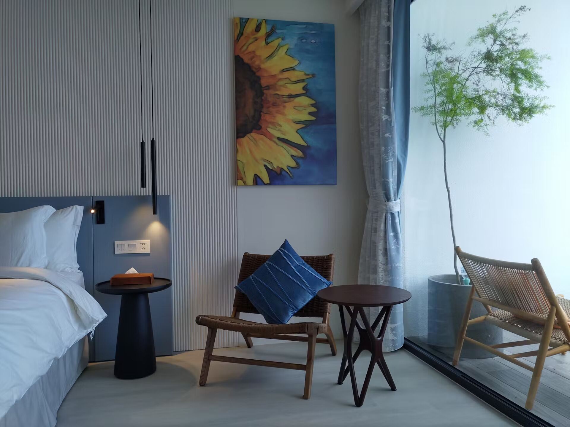 A hotel room with a bed, a small table, a chair, a painting, and a window with a plant.
