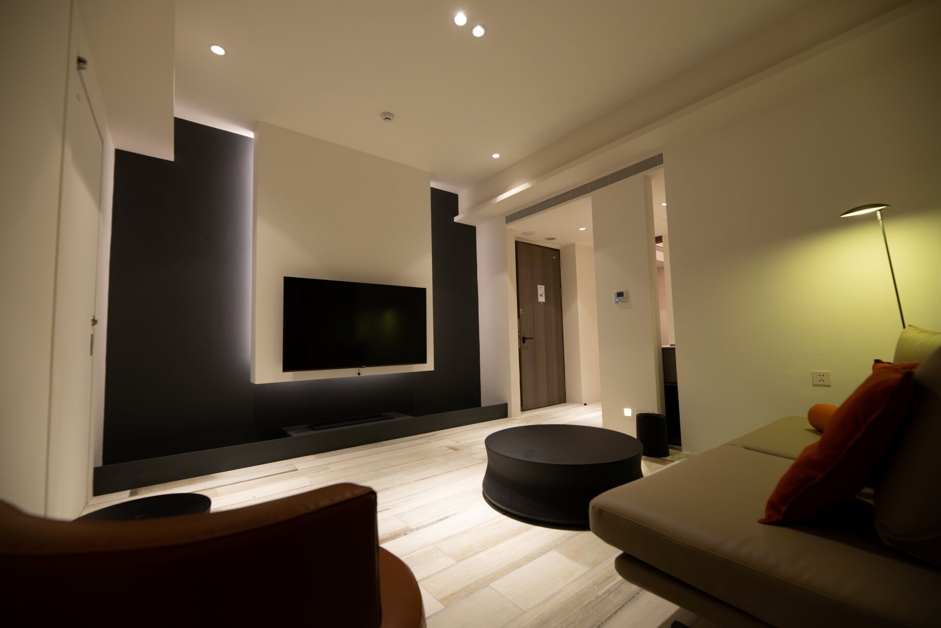 A modern living room with a flat-screen TV mounted on the wall, a round black ottoman, and a sofa with cushions.