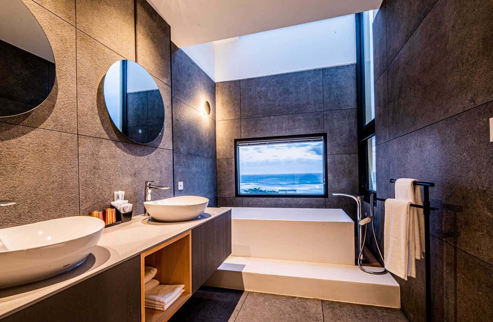 A modern bathroom with a large bathtub, a round mirror, and a window with a view of the ocean.