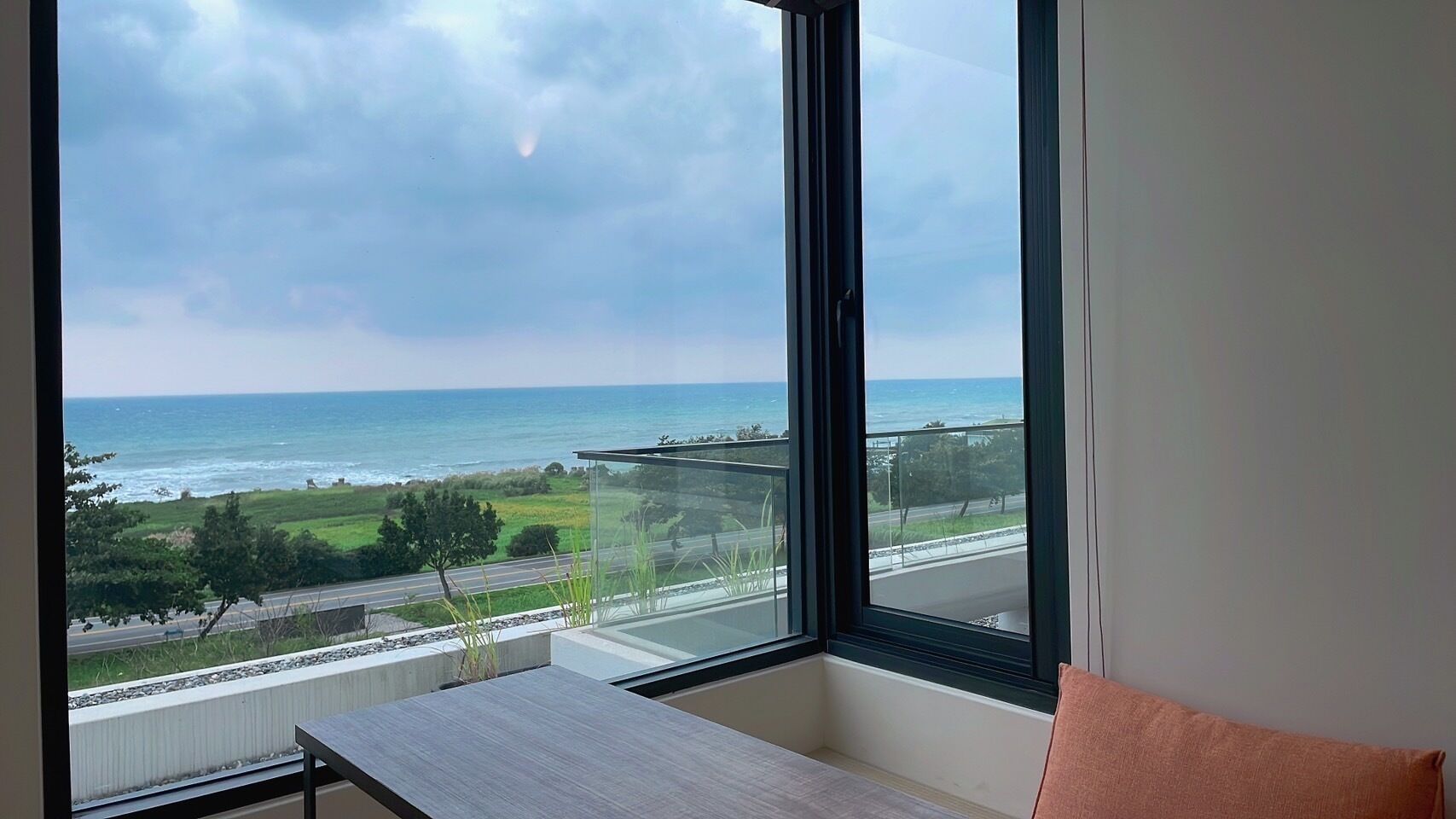 A room with a large window overlooking a coastal view, a small table, and a cushion.