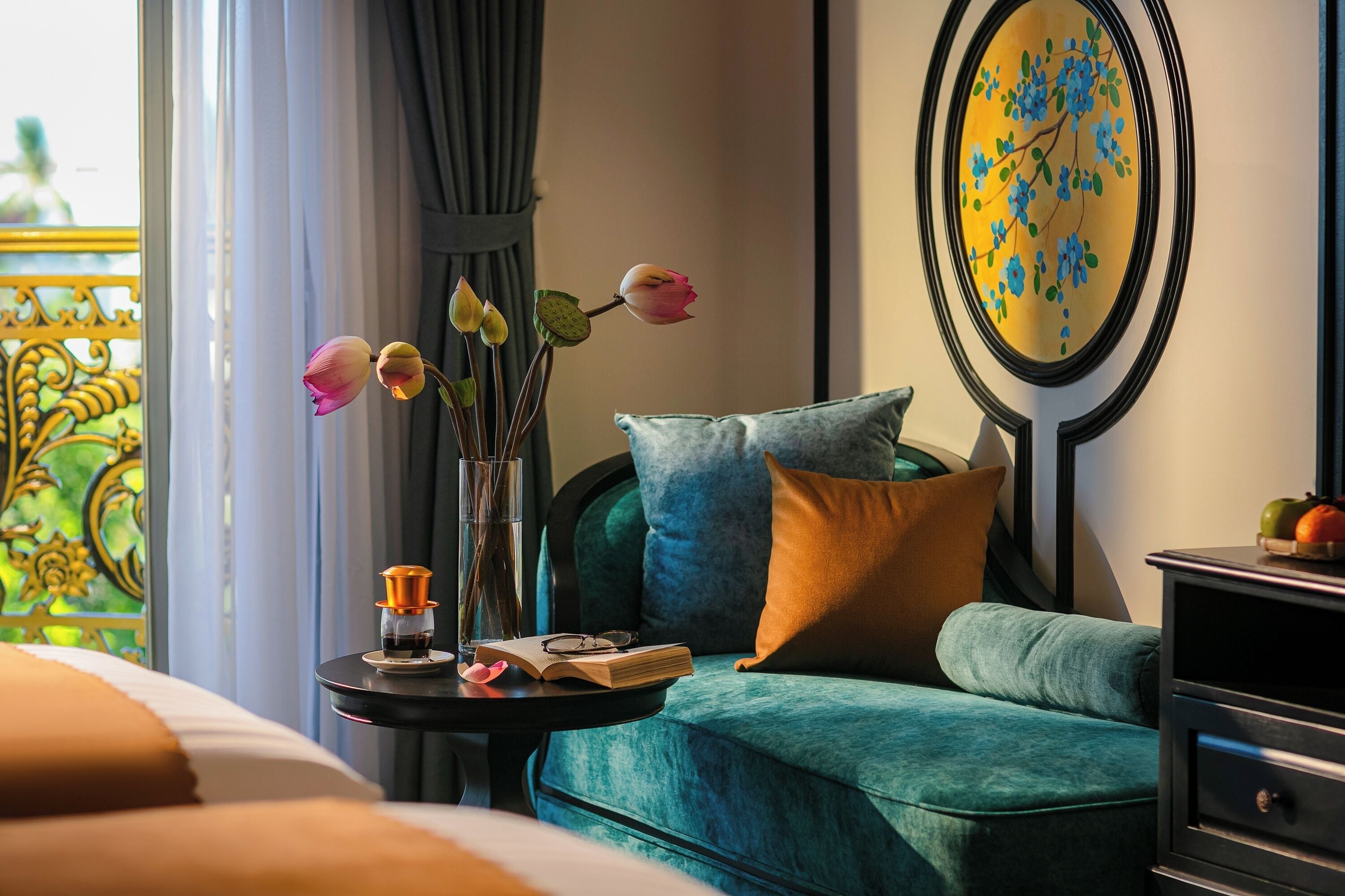 Una habitación de hotel con una cama, un sofá color turquesa, una mesita con un jarrón de flores y un cuadro en la pared.
