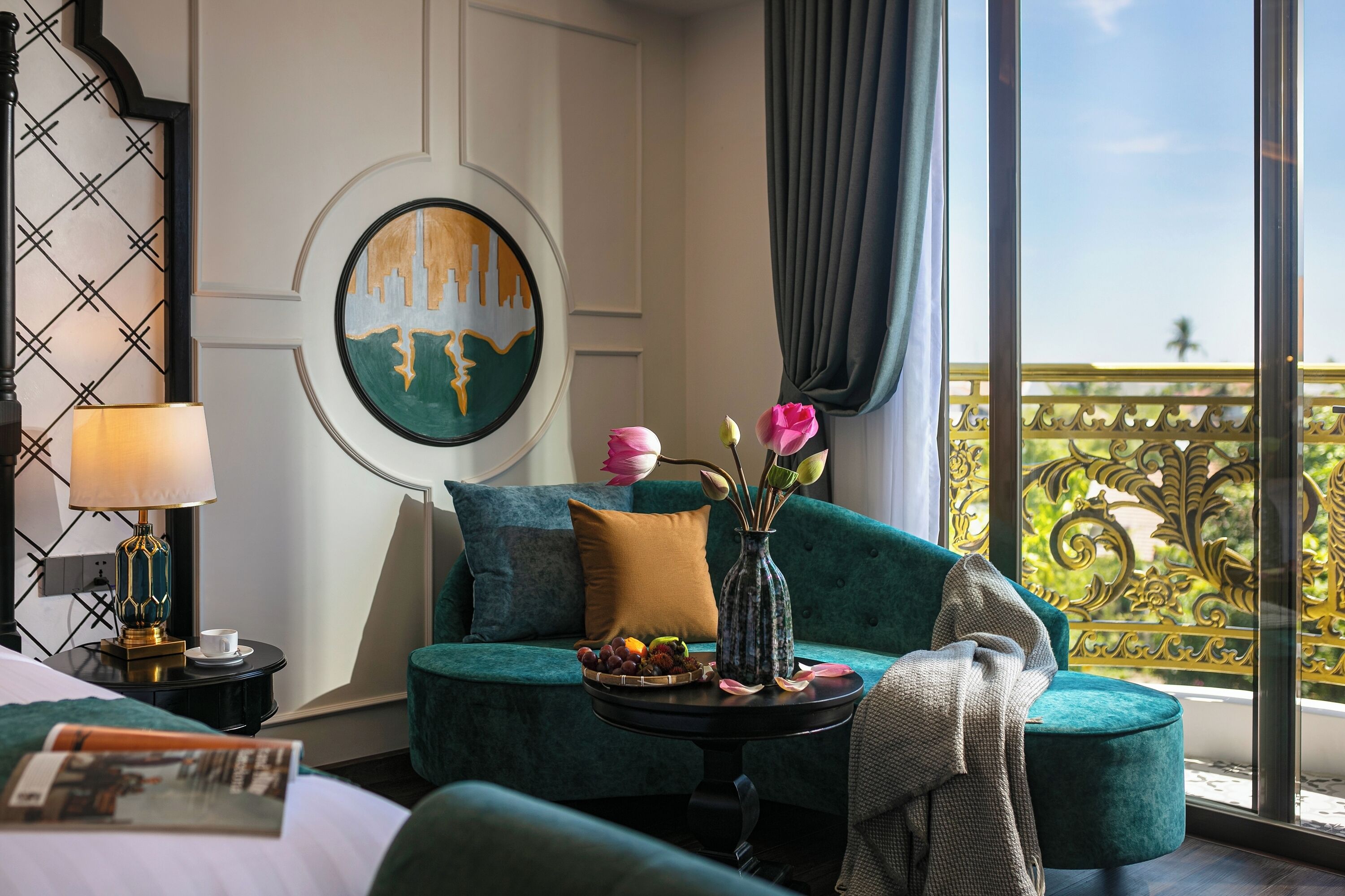 Quarto de hotel com sofá azul-turquesa, uma pequena mesa redonda com um vaso de flores, um abajur e um quadro na parede.