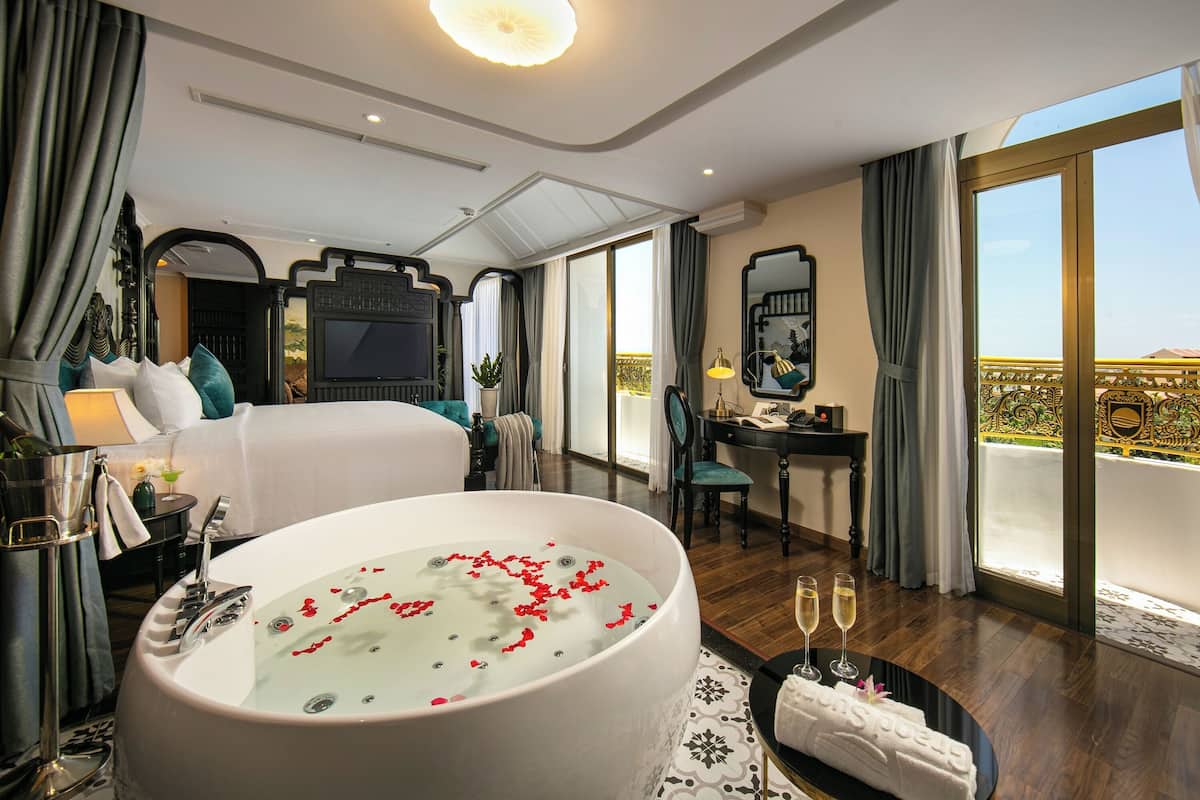 Une chambre d’hôtel luxueuse avec un grand lit, une baignoire remplie de pétales de roses, un coin repas et un balcon offrant une vue imprenable.