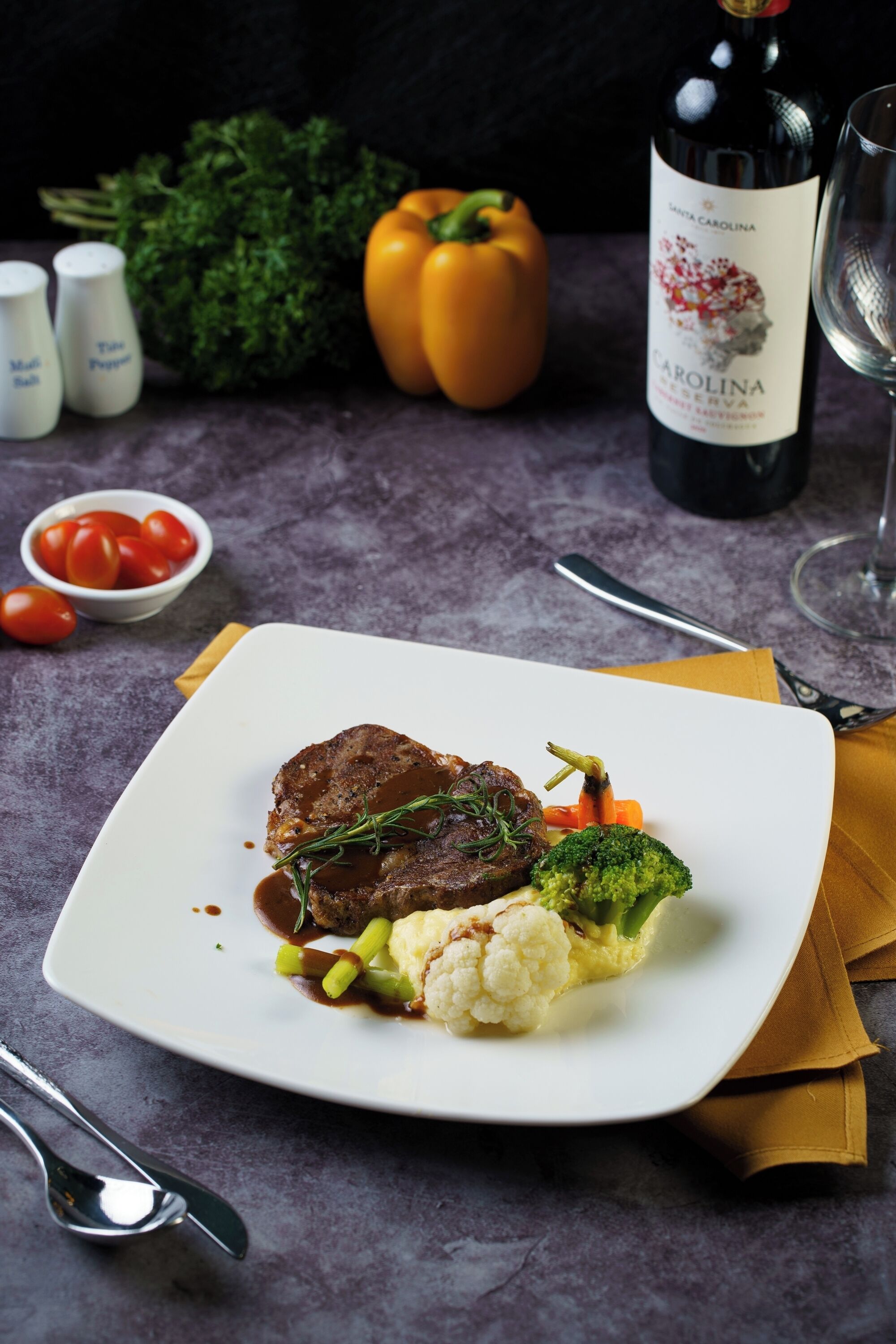 A plate of food with steak, vegetables, and sauce, accompanied by a bottle of wine and a glass on a table.