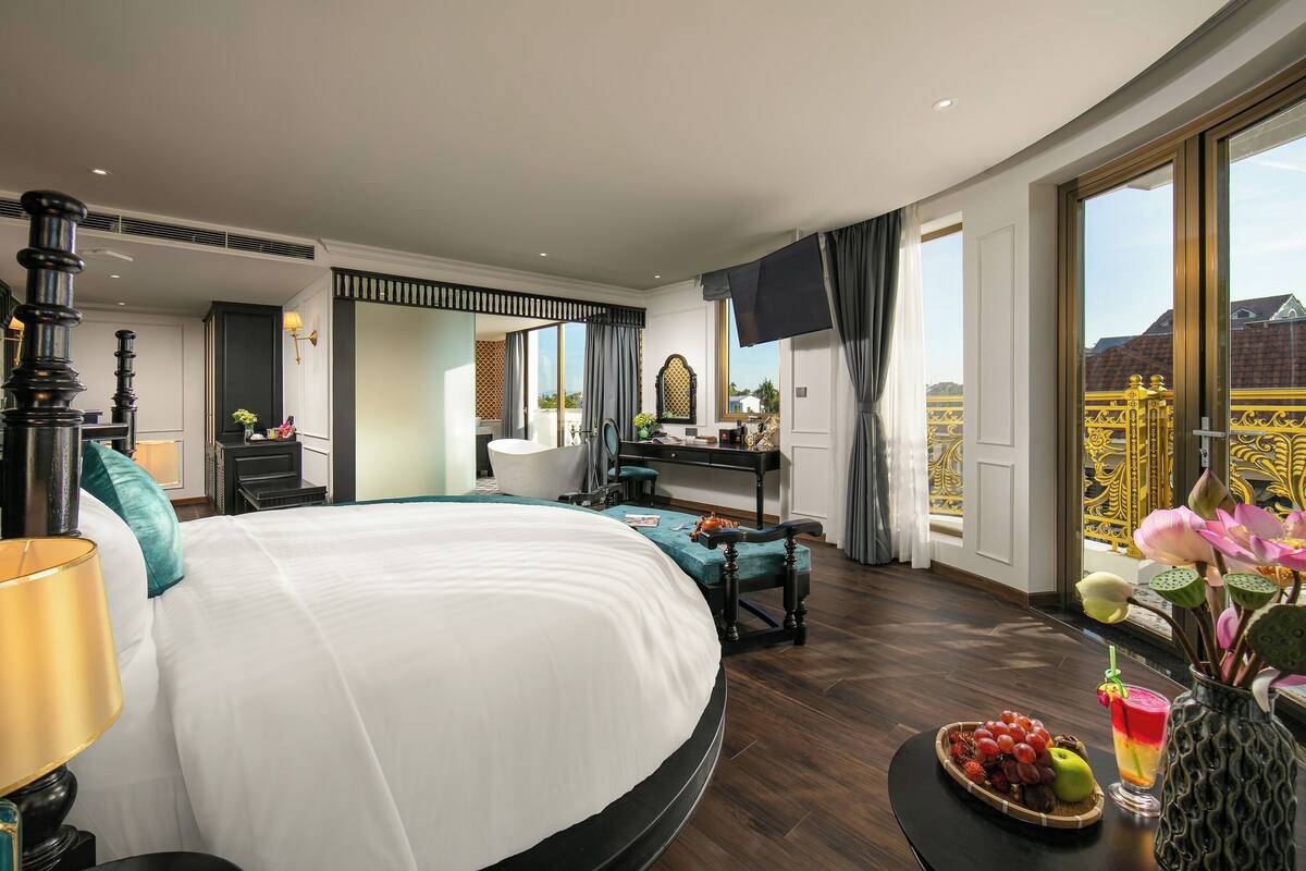 Une chambre d’hôtel spacieuse, dotée d’un grand lit, d’un bureau, d’une chaise et d’une table à manger avec des fruits et une boisson.