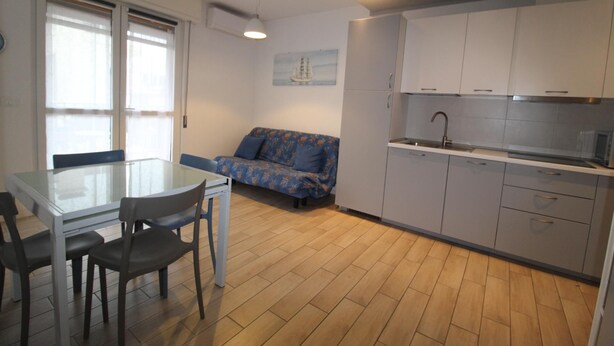 A compact kitchen with grey cabinets, a white countertop, and a sink. There is a dining table with chairs and a sofa with a floral pattern. - Alideco - Lignano Sabbiadoro (Lignano Sabbiadoro)