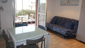 A small, well-lit room with a glass table and chairs, a sofa, and a painting of a sailboat on the wall. - Alideco - Lignano Sabbiadoro (Lignano Sabbiadoro)