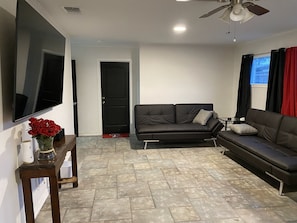 A living room with a black leather sofa, a flat-screen TV mounted on the wall, and a ceiling fan. - Our Pool House (Lubbock)