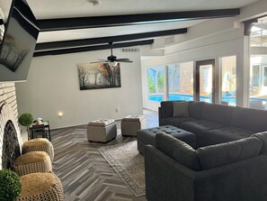 A modern living room with a sectional sofa, ottomans, a herringbone-patterned floor, a painting, and a view of a swimming pool. - Our Pool House - Swim Rain or Shine (Lubbock)
