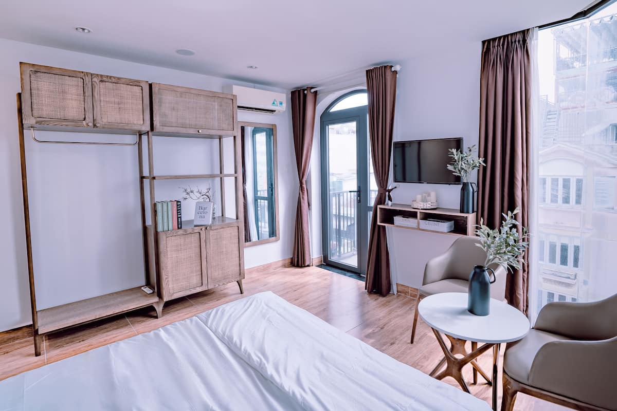 Habitación de hotel moderna con una cama grande, un escritorio con televisor, una zona de estar con mesa y sillas, y un balcón con cortinas.