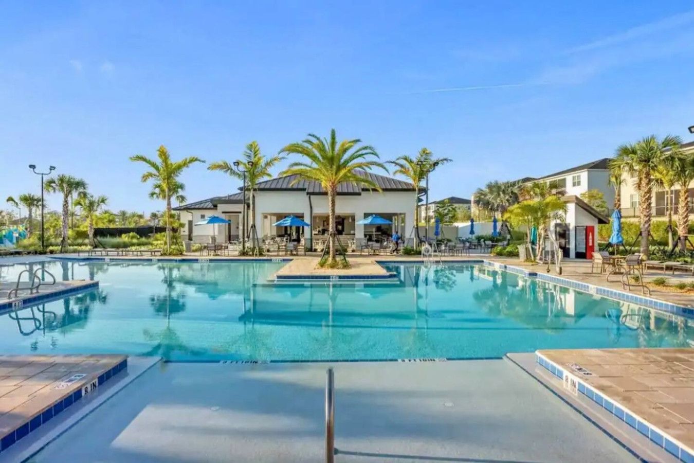A swimming pool with lounge chairs and umbrellas, surrounded by palm trees and buildings.