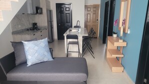 Un espacio habitable compacto con sofá, una mesa de comedor con sillas, una zona de cocina y un pequeño estante de madera.