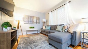A living room with a grey sofa, a coffee table, a TV mounted on the wall, a small side table with a lamp, and a large window with curtains. - Simple & Sweet 2 BR 1 BA Home in Lansing - Cynwood (Lansing)