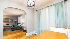 A room with a wooden floor, a bed with a white headboard, a small table, a lamp, and a view through an archway to another room with a sofa and a painting. - Simple & Sweet 2 BR 1 BA Home in Lansing - Cynwood (Lansing)