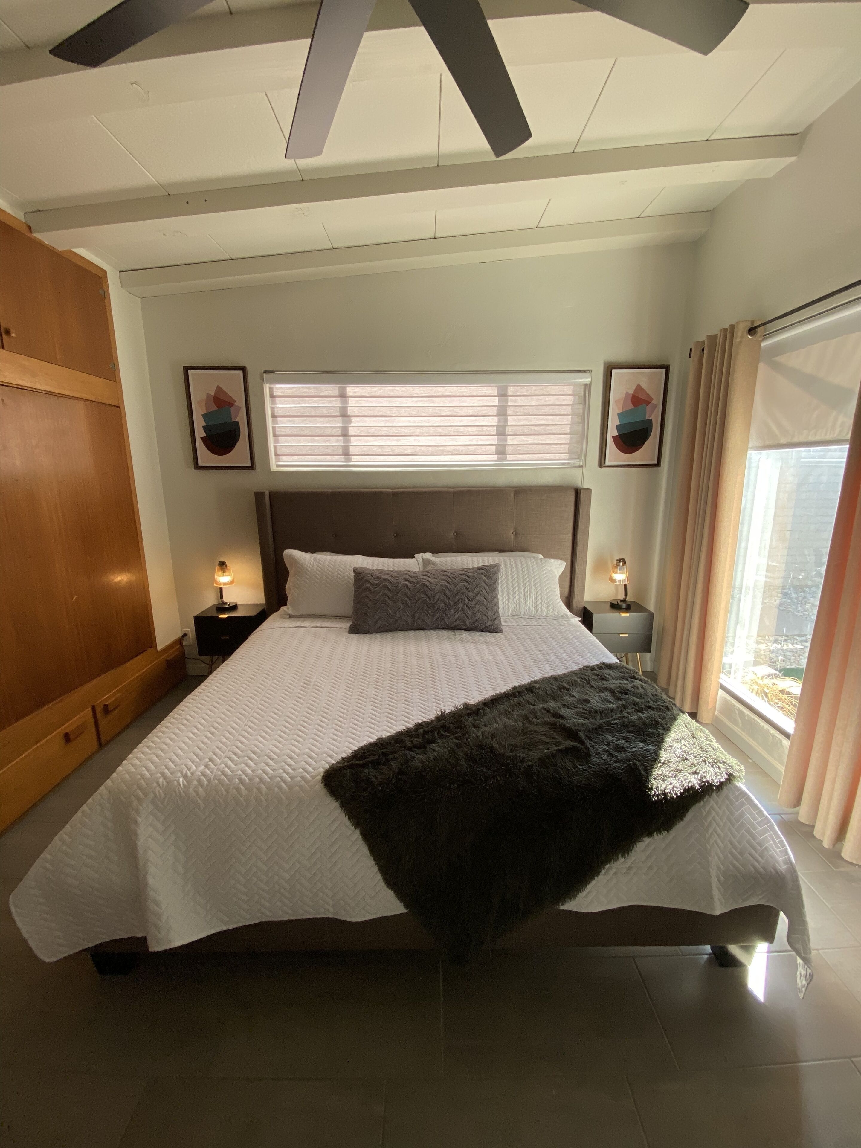 A bedroom with a large bed, wooden wardrobe, two bedside tables with lamps, a window with curtains, and artwork on the walls.