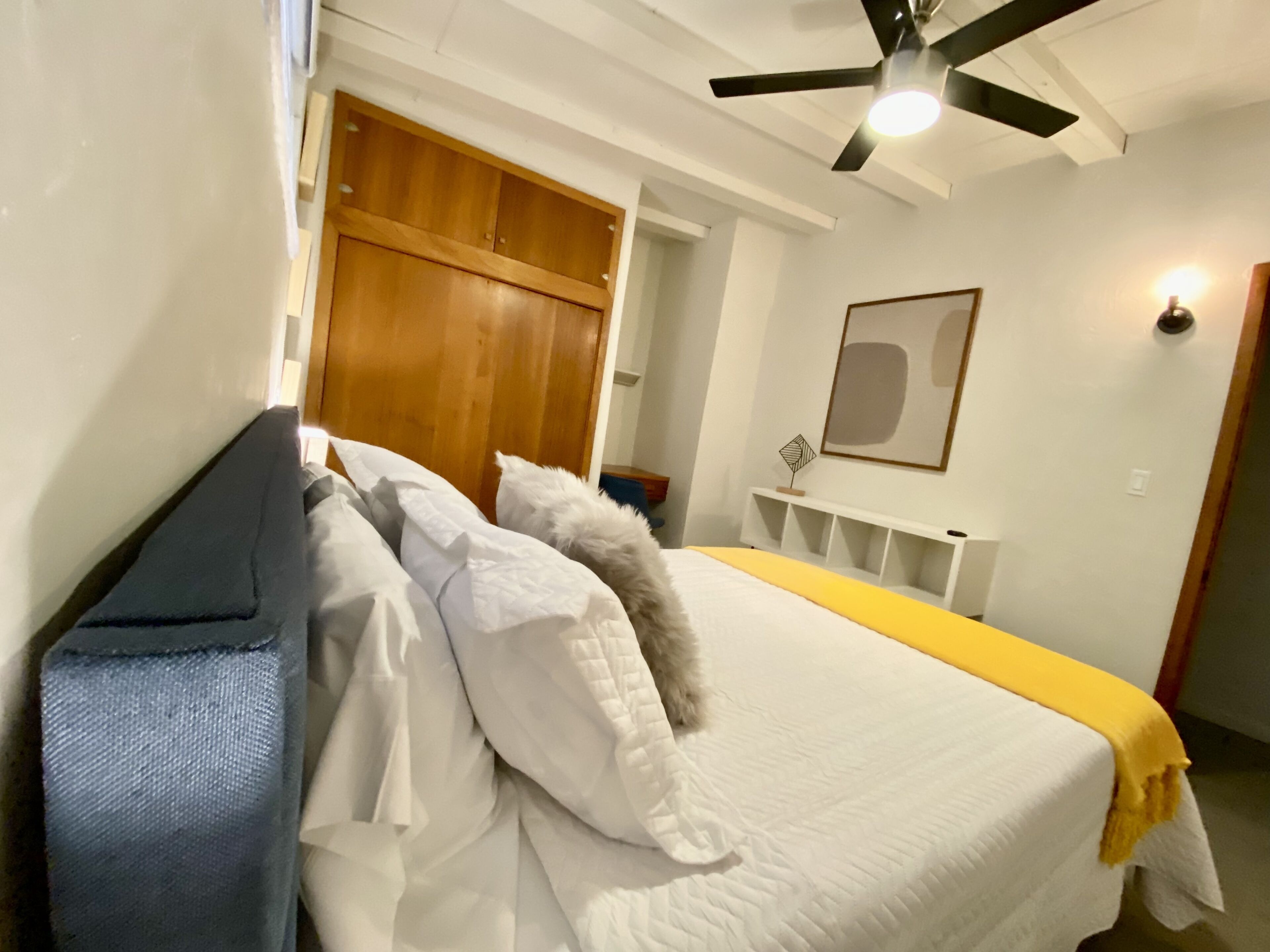 Un lit bien fait, avec du linge de lit blanc et un plaid jaune, une armoire en bois, un ventilateur de plafond et un tableau encadré au mur.