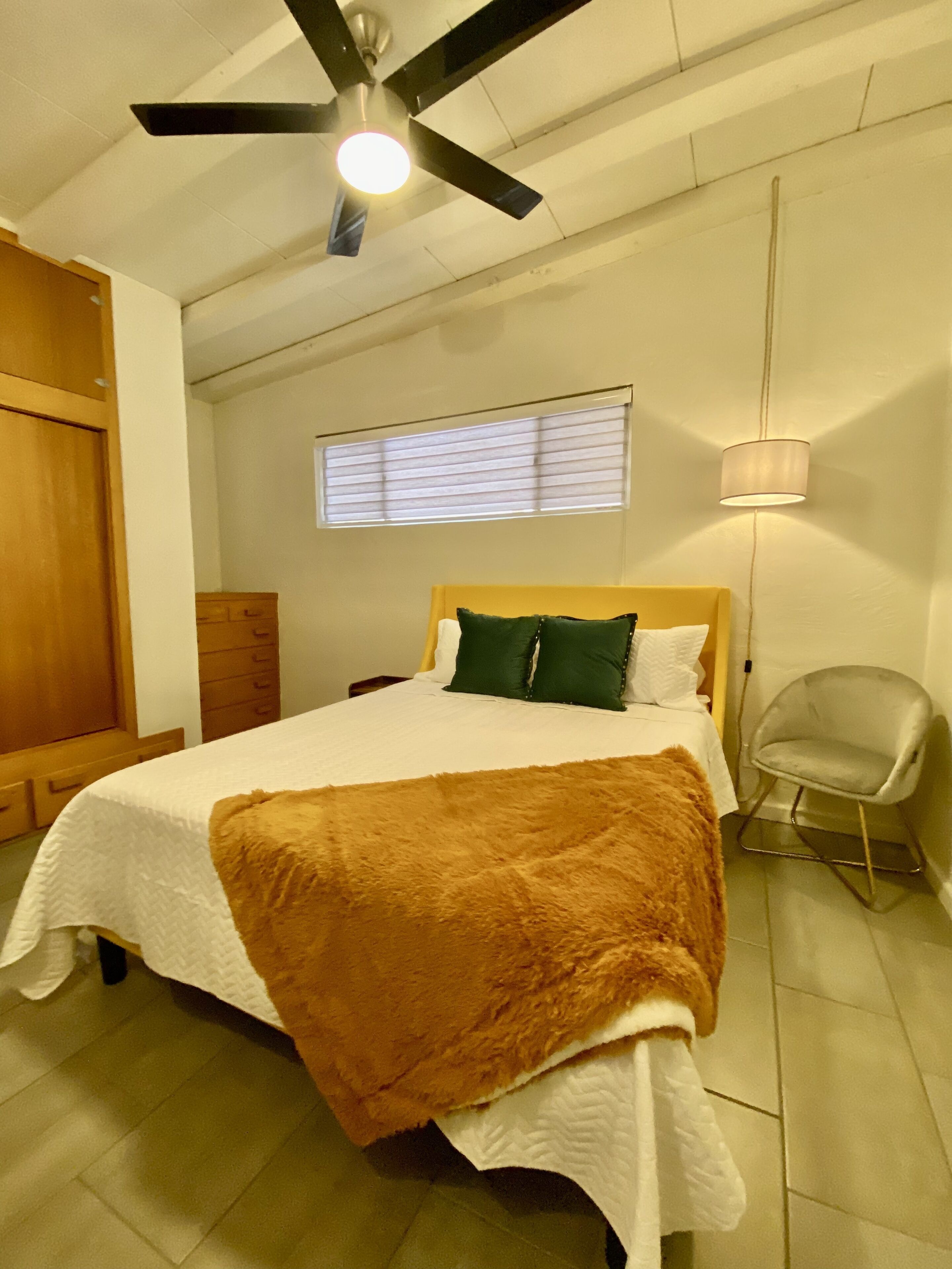 Une chambre à coucher comprenant un lit, une fenêtre avec des stores, un ventilateur de plafond, une chaise et une armoire.