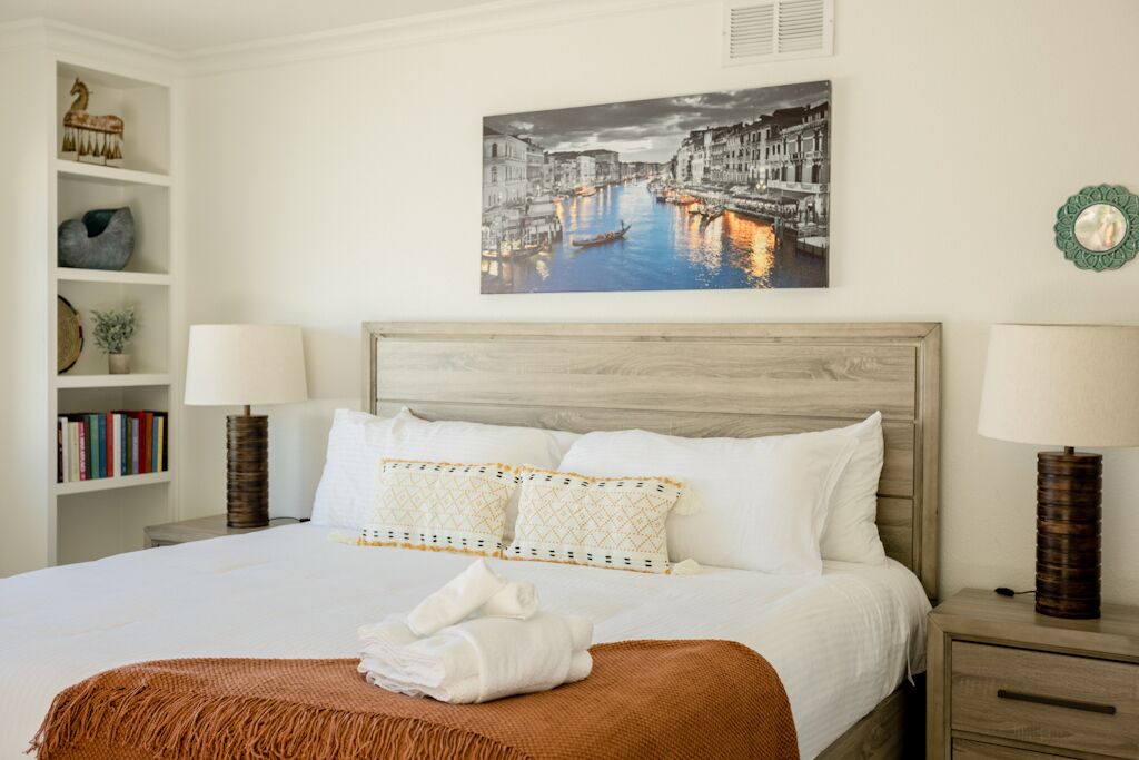 Una cama bien tendida con sábanas blancas y almohadones estampados, un cabecero de madera y un cuadro enmarcado con una vista urbana.