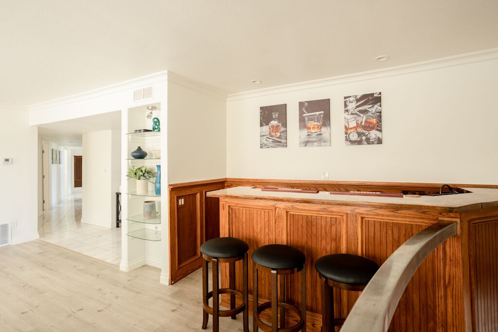 A kitchen with a wooden bar and stools, a glass shelf with decorative items, and wall art depicting drinks.